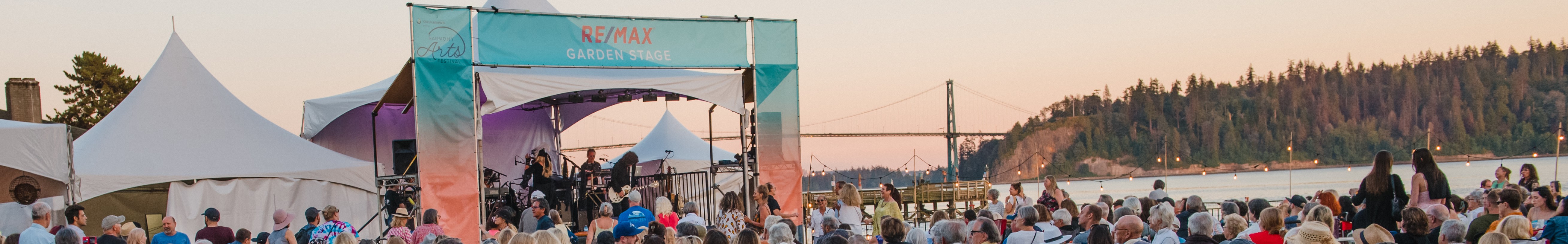 A crowd of people standing in front of tents at Re/Max garden stage with ocean and lions gate bridge in background.