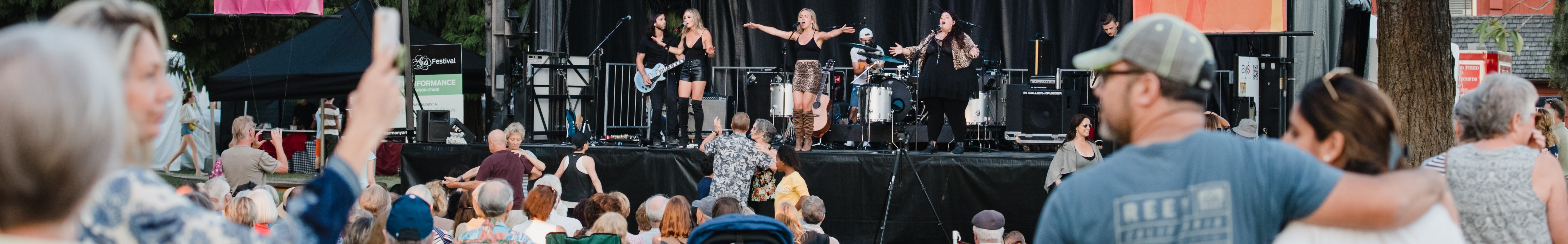 The image is a photo from the Harmony Arts Festival performance featuring a blond woman with a microphone on stage and a crowd of people. The scene captures a live music event with a mix of men and women standing outdoors enjoying the performance